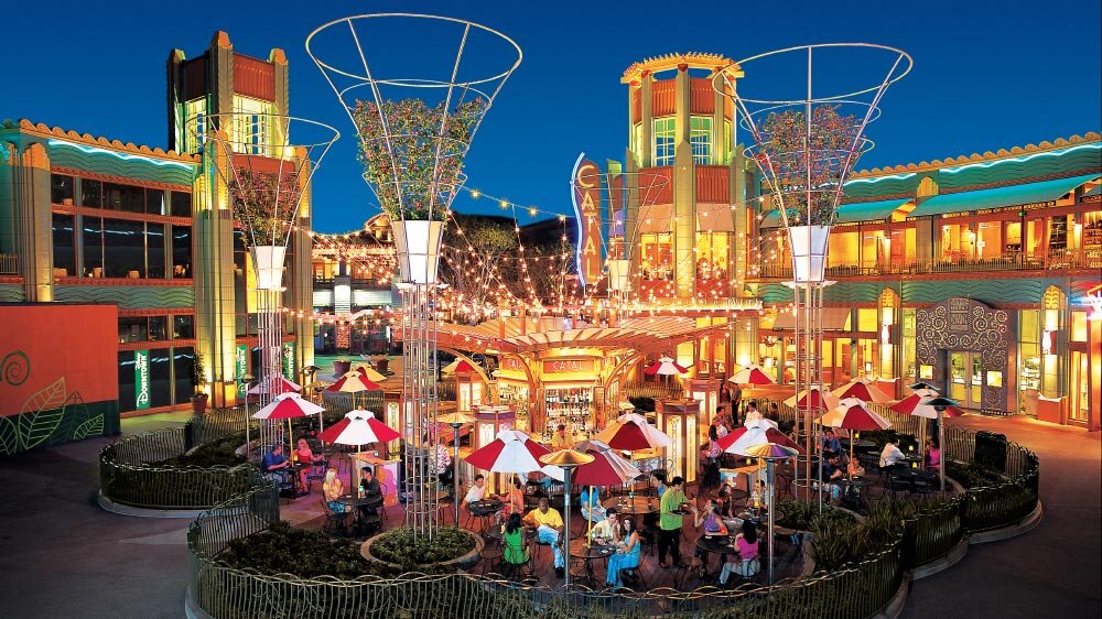 A vibrant outdoor seating area at night with people sitting at tables under colorful umbrellas, surrounded by decorative lighting and buildings.