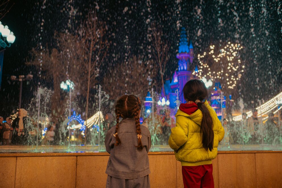 Two girls looking at a fountain and castle at night with snow falling.